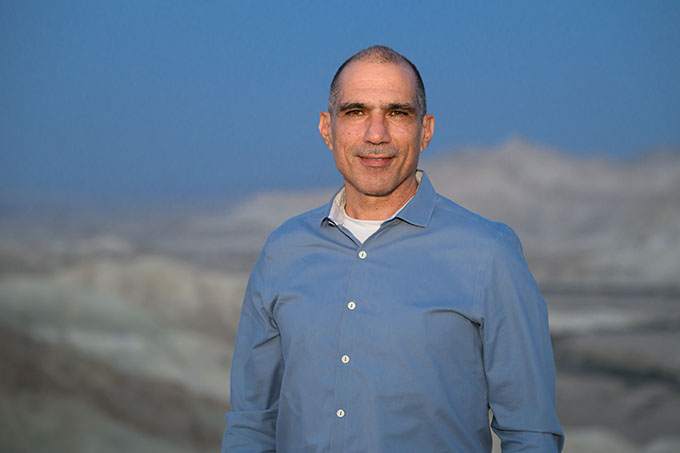 The image shows a middle-aged man wearing a blue shirt in a mountainous landscape.